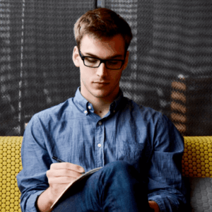 A young man in glasses and a blue shirt sits on a yellow couch, focused on writing in a notebook, with a modern, textured background