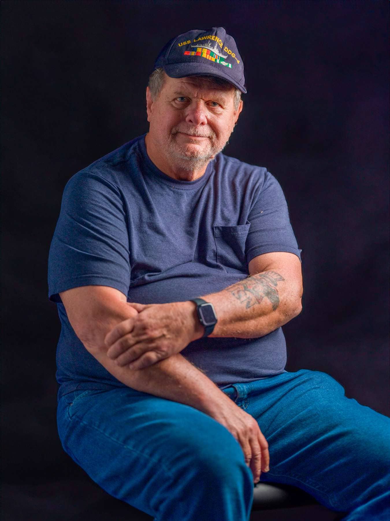 Man wearing USS Lawrence DDG cap, blue t-shirt, and jeans, posing for professional portrait.